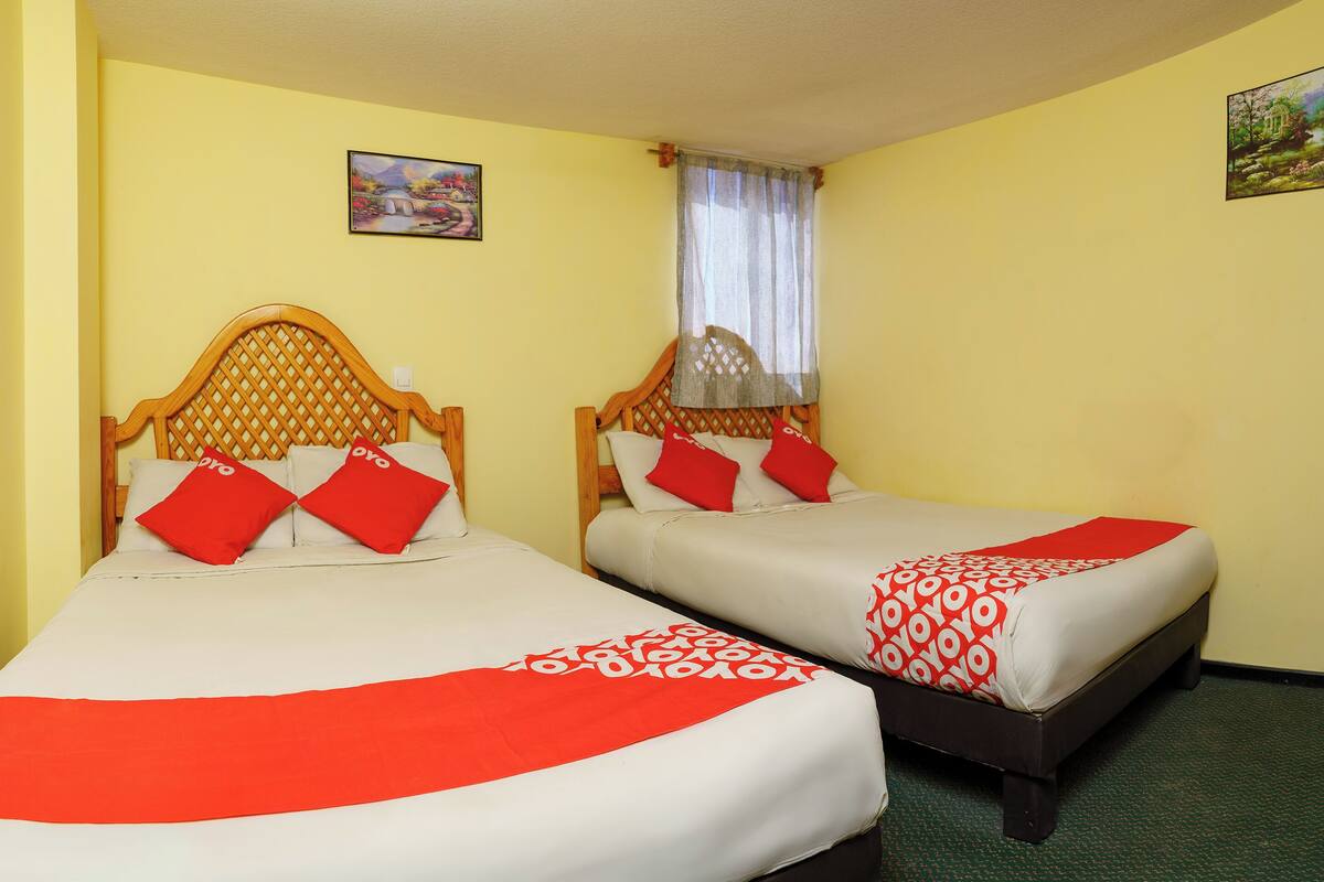 Dos camas individuales con cabeceros de madera, ropa de cama roja y blanca, y una pared amarilla con un cuadro.