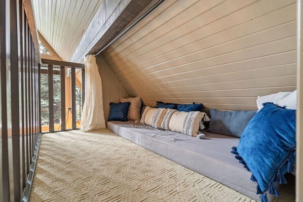 Un dormitorio acogedor en el ático, con techo inclinado, un banco empotrado con cojines y ventanas grandes.
