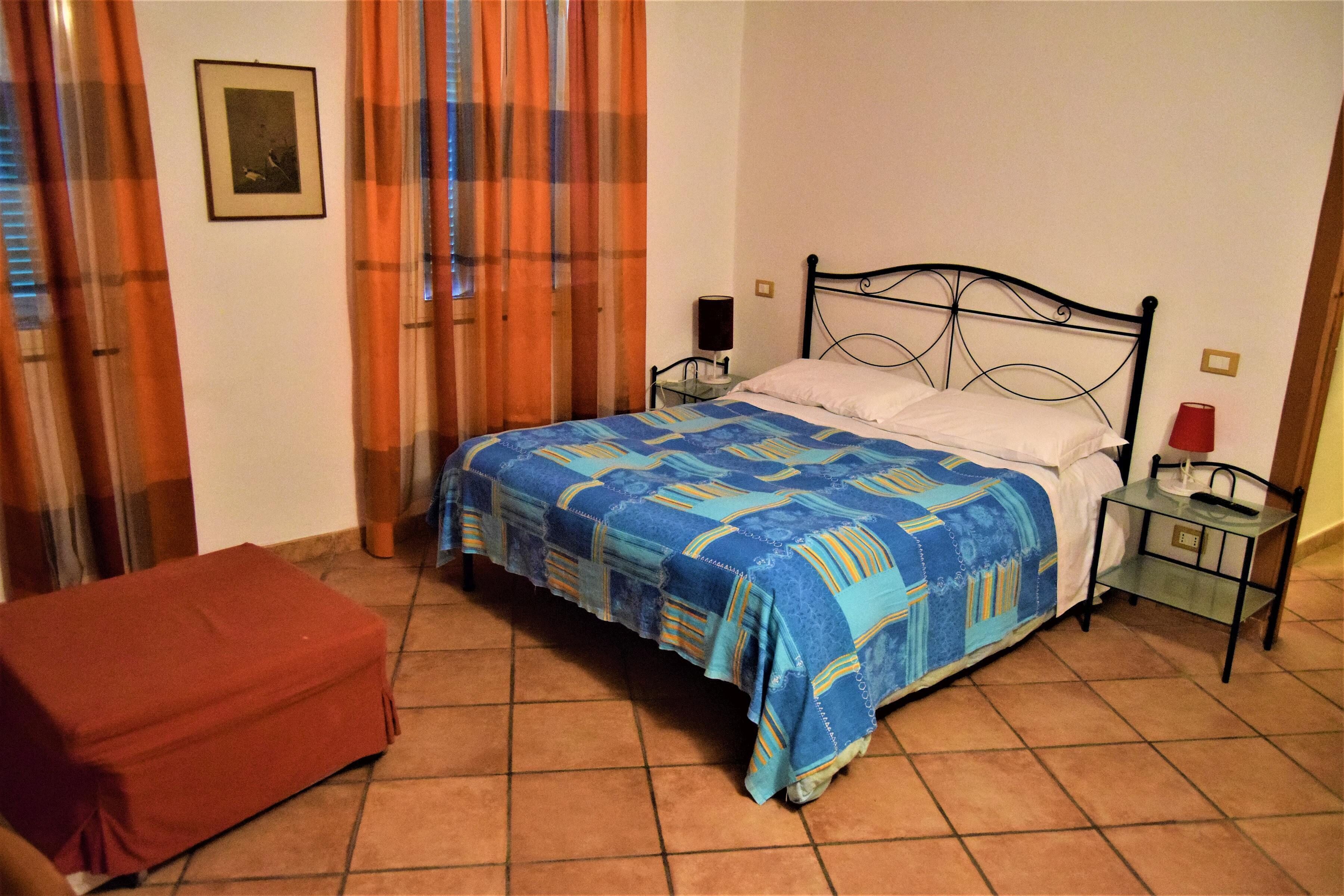 Un dormitorio con una cama, una mesita de noche, un reposapiés rojo, una ventana con cortinas y un cuadro colgado en la pared.