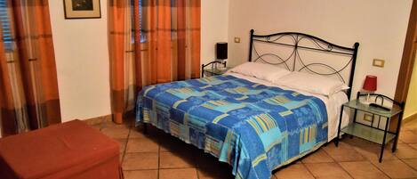 Un dormitorio con una cama, una mesita de noche, un reposapiés rojo, una ventana con cortinas y un cuadro colgado en la pared.