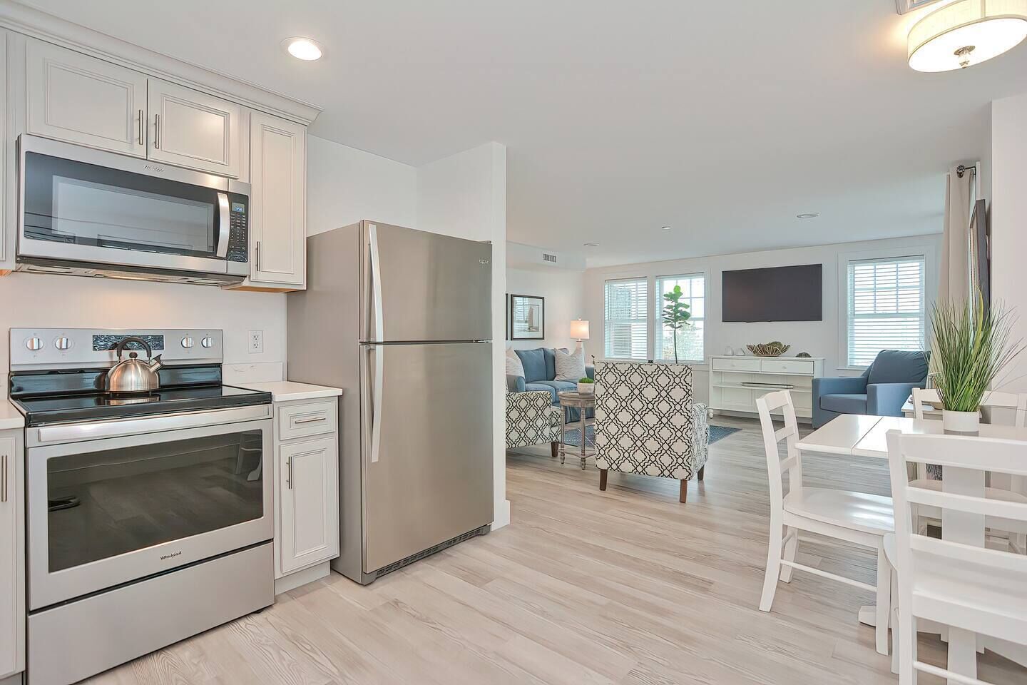 A modern kitchen with stainless steel appliances, a dining area with white furniture, and a living room with a TV and seating.