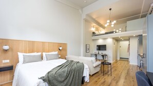 A modern hotel room with a wooden floor, a large bed with white bedding, a grey sofa, a small table with a lamp, and a TV mounted on the wall. - Foundry Hotel Apartments (Uppsala)