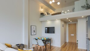 A modern living room with a sofa, dining table, and chairs. There is a TV mounted on the wall, and a wooden floor. - Foundry Hotel Apartments (Uppsala)