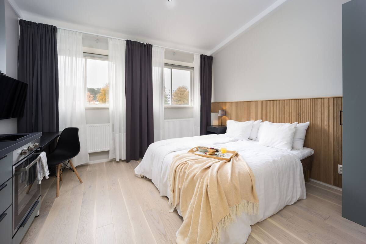 A modern hotel room with a bed, a desk with a chair, a television, and a window with curtains.
