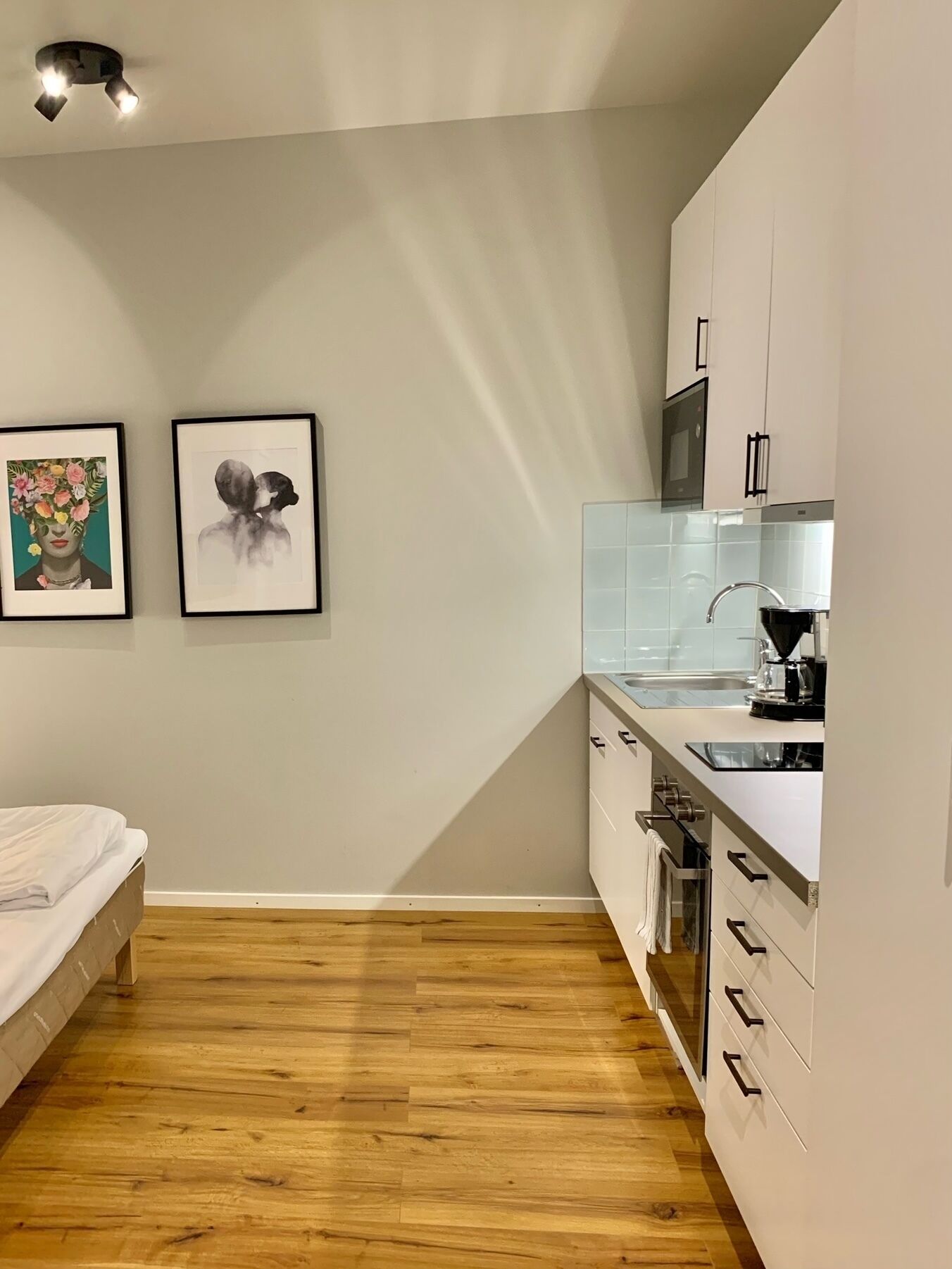 A compact kitchen with white cabinets, a stainless steel sink, and a microwave. There is a bed with a white sheet and a wooden floor. Two framed artworks are hung on the wall.