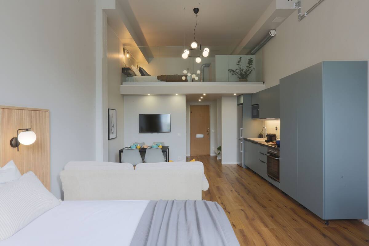 A modern hotel room with a large bed, a dining area, a kitchen, and a balcony.
