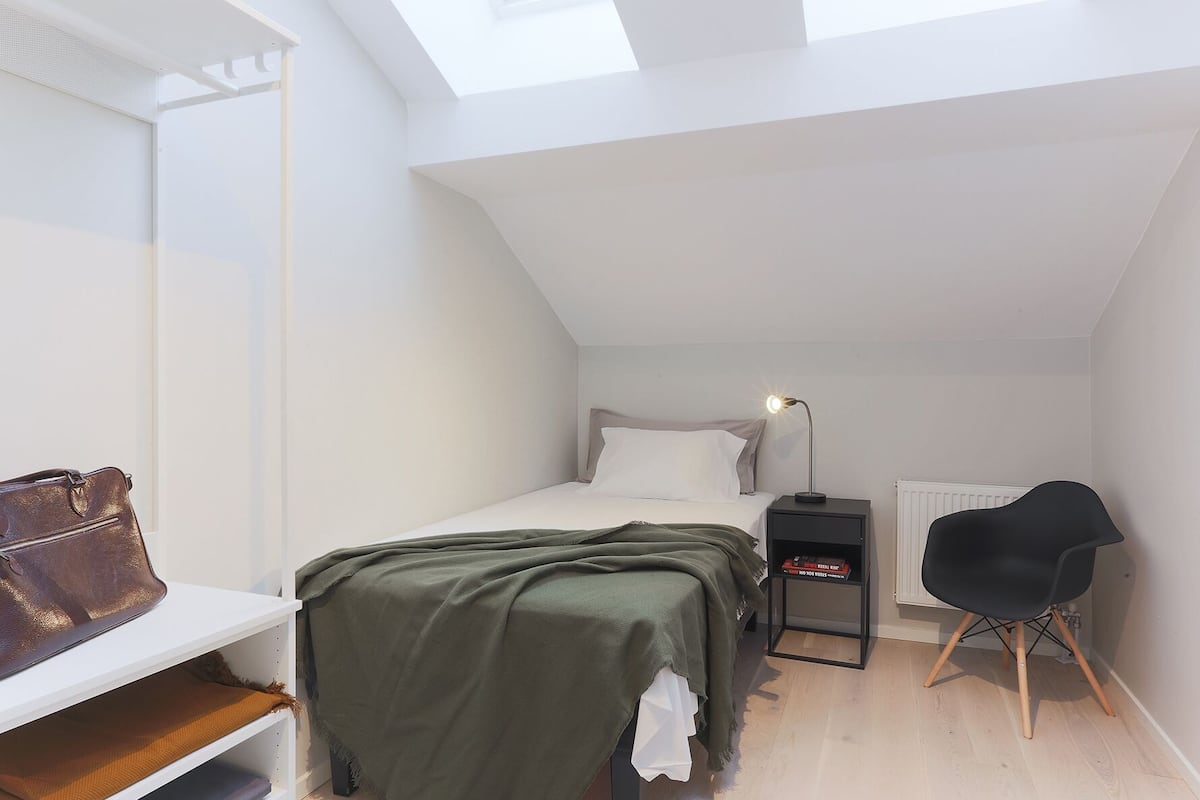 A small, modern bedroom with a sloped ceiling, a bed, a bedside table with a lamp, a chair, and a radiator.