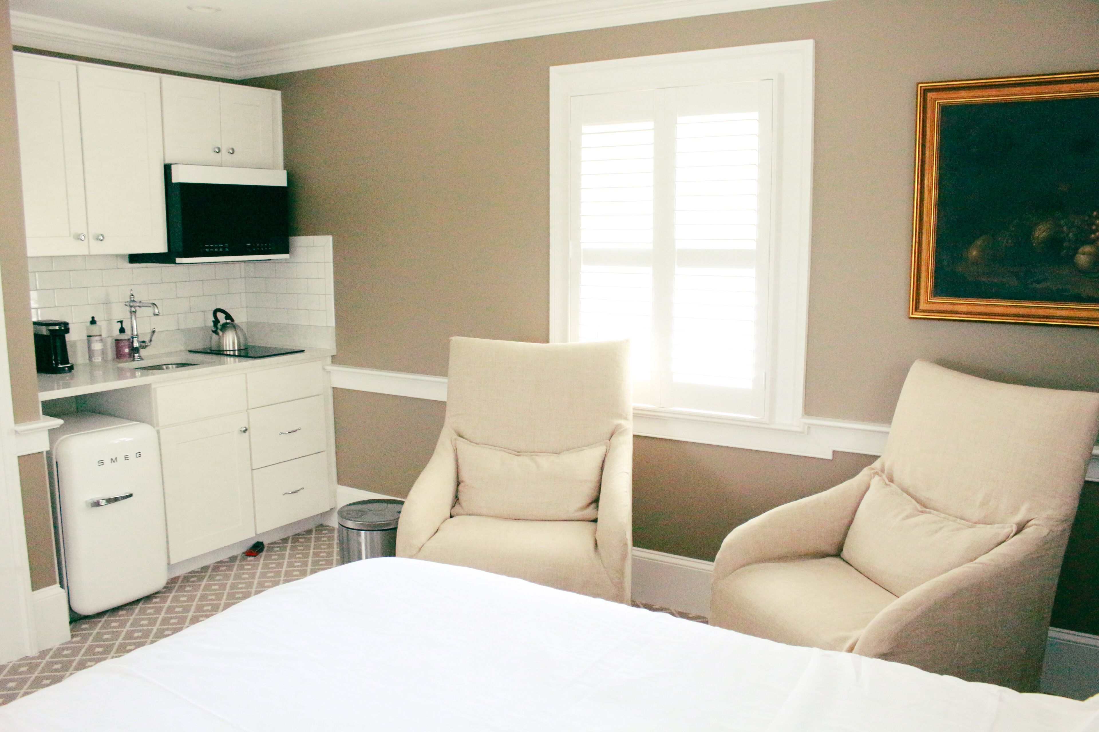 A bedroom with a bed, two armchairs, a kitchenette, and a window.