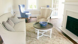A living room with a beige sofa, a blue armchair, a round coffee table with a blue bowl, a fireplace, and a window with a floral valance. - Inn at Mary's Land Farm (Clarksville)