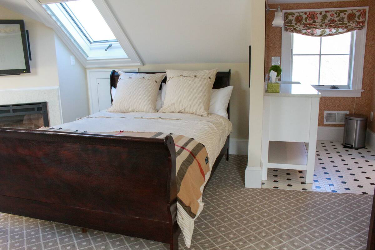 A bedroom with a bed, a skylight, a fireplace, a window with curtains, and a small kitchenette area.