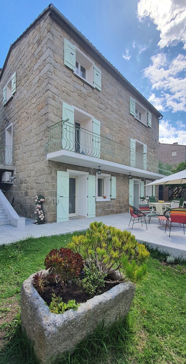 A stone building with a balcony, a garden with plants, and outdoor furniture.