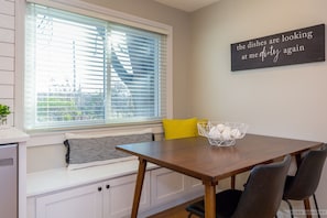 Un coin repas avec une table en bois, des chaises noires et jaunes, un banc blanc agrémenté d’un coussin à carreaux et une fenêtre avec des stores.