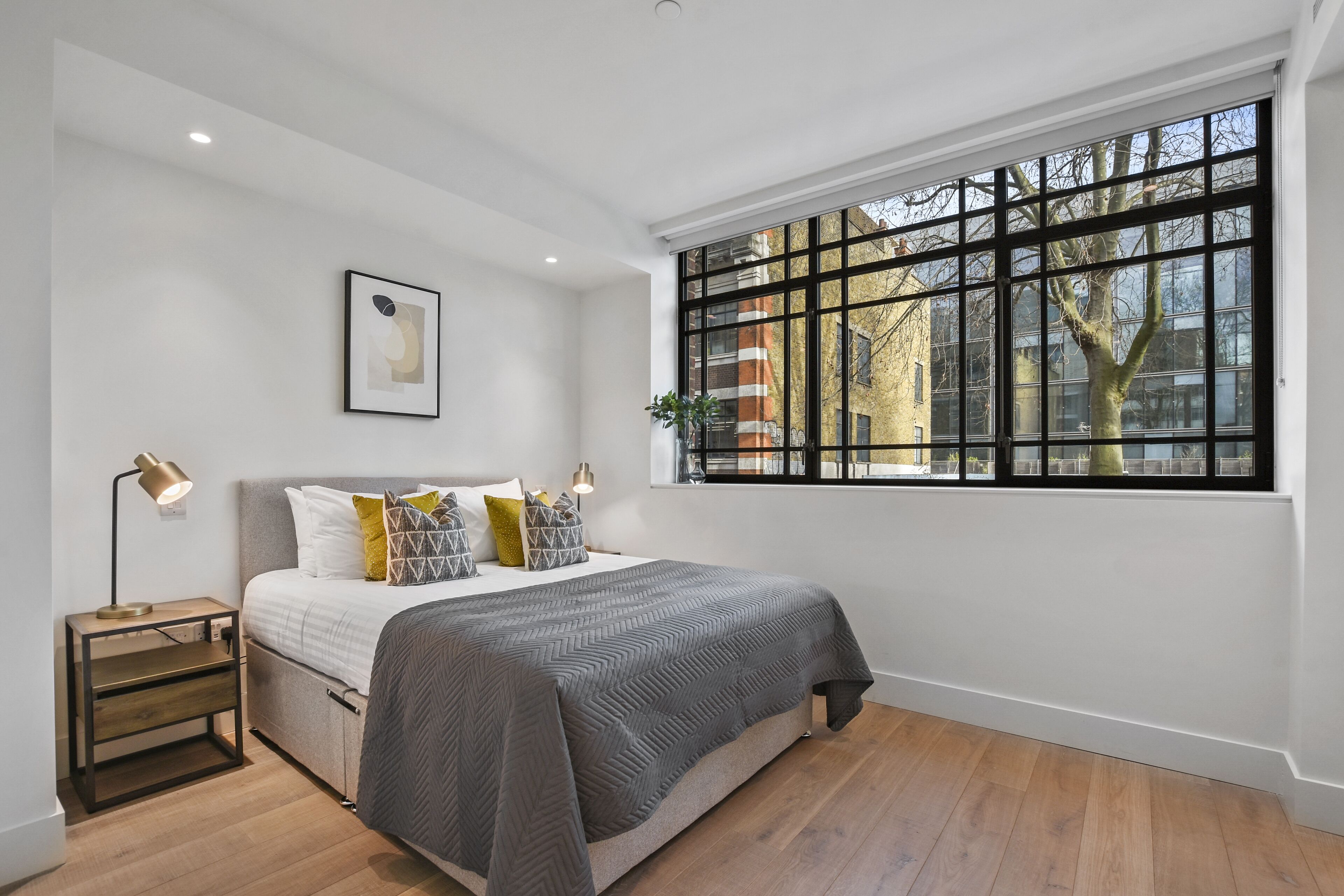 A bedroom with a large window, a bed with a grey quilt, a bedside lamp, and a framed picture on the wall.