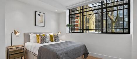 A bedroom with a large window, a bed with a grey quilt, a bedside lamp, and a framed picture on the wall.
