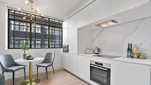 A modern kitchen with a round dining table, two chairs, a built-in oven, and a window with a view of buildings.