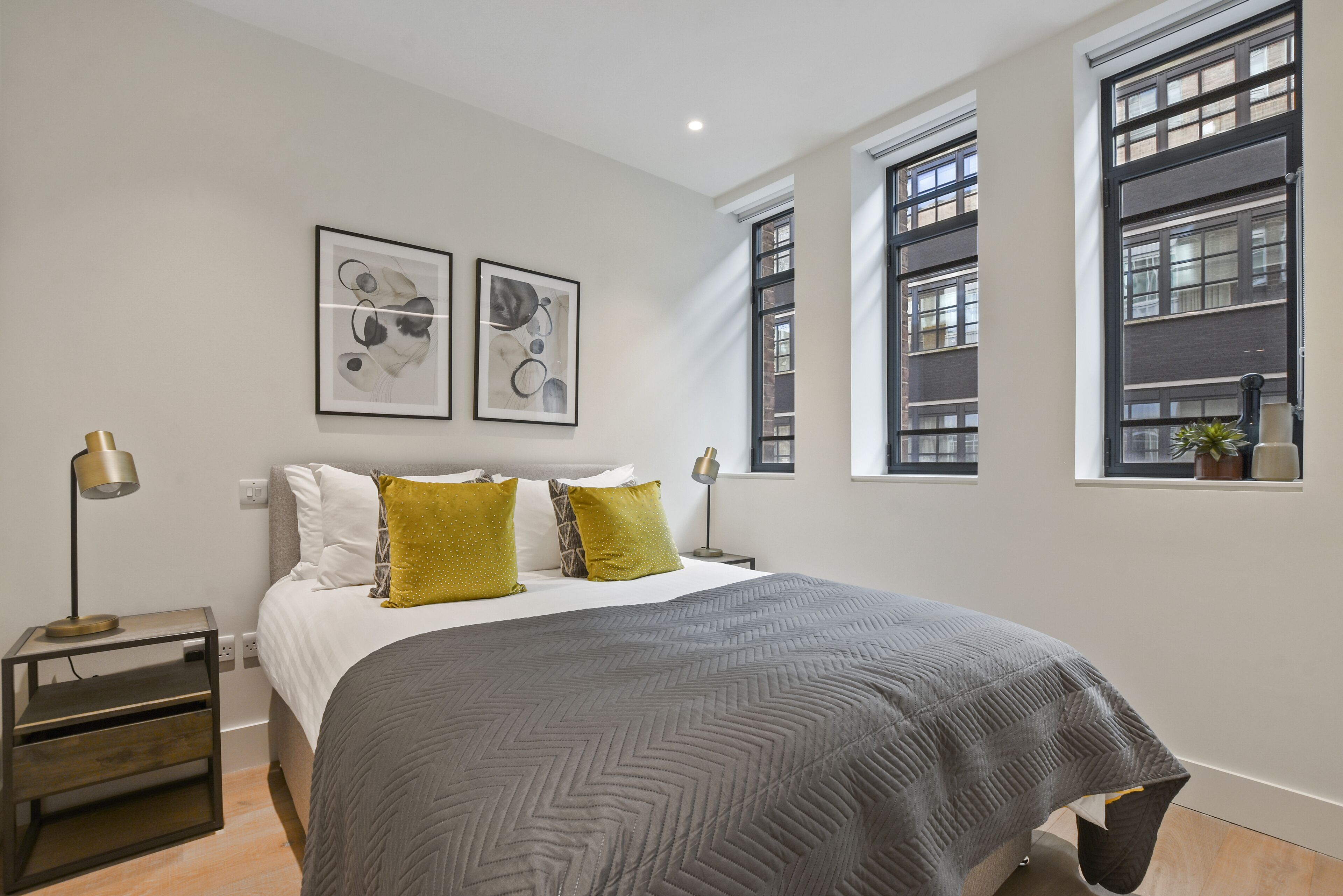 A modern bedroom with a large bed, two side tables, and two windows with views of a building.