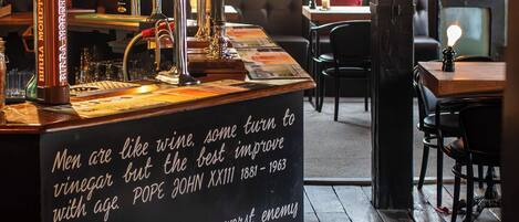A bar area with a chalkboard displaying text, a variety of bottles, glasses, and a seating area with a large window.