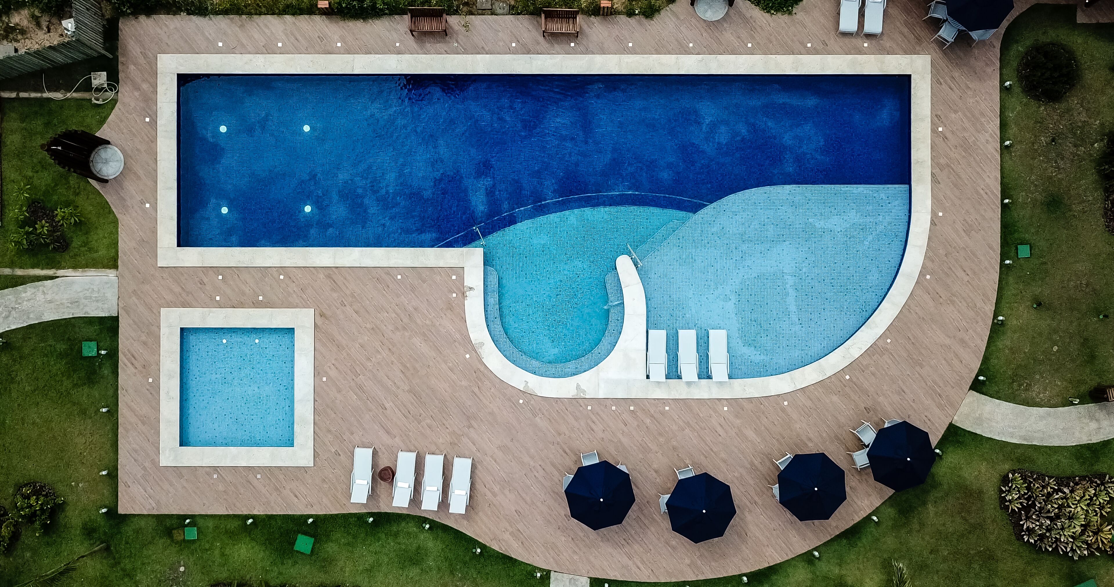 Vista aerea di una zona piscina con una piscina rettangolare, una piscina più piccola e un’area relax con ombrelloni.