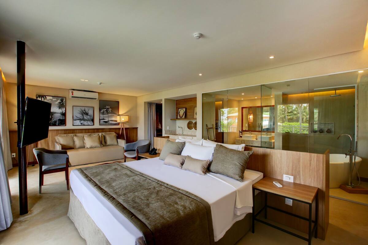 A modern hotel room with a large bed, a sitting area with a sofa and chairs, a TV, and a bathroom with a glass shower enclosure.