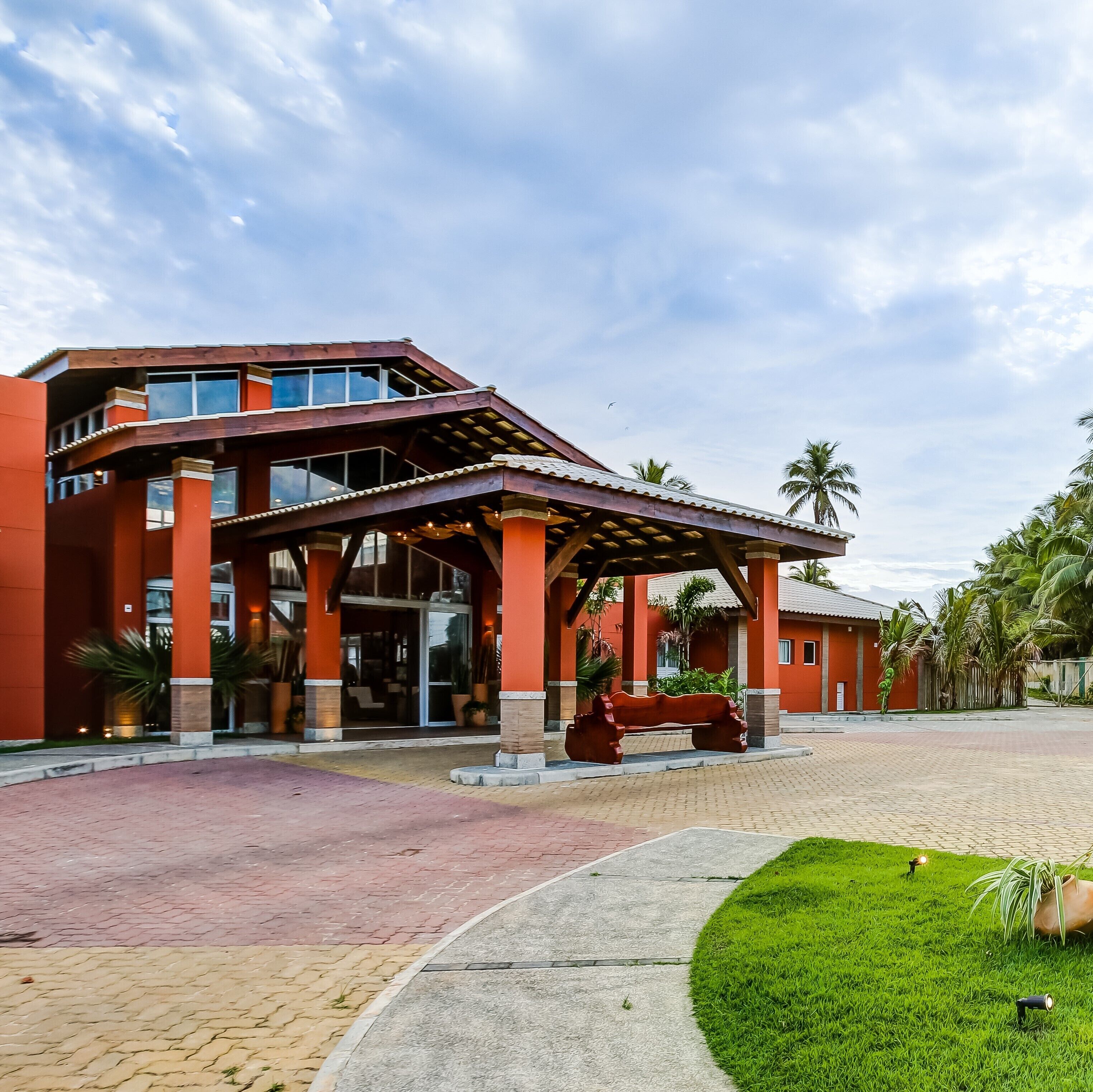 Un edificio moderno con entrada cubierta, palmeras y un área pavimentada con un banco.