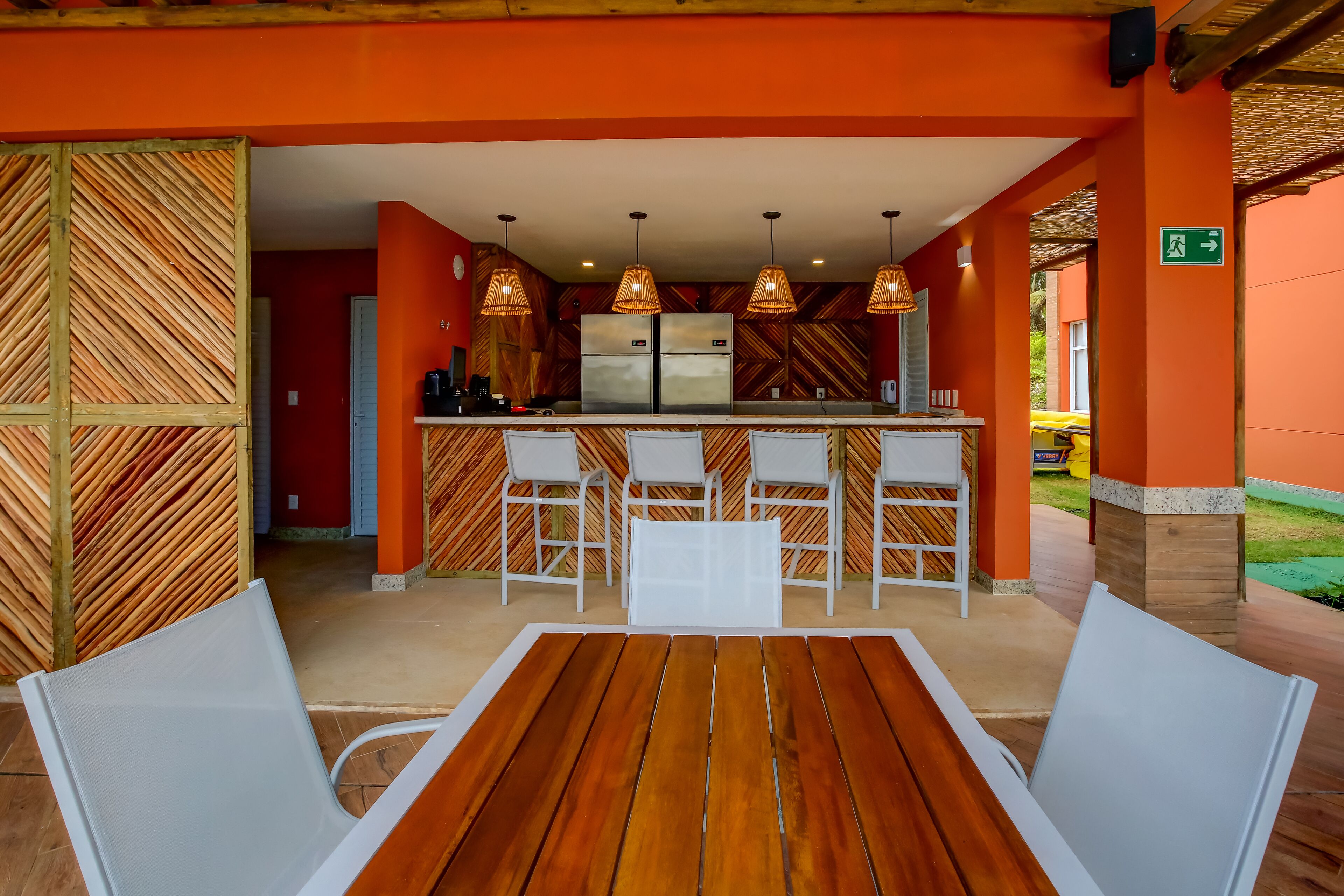 A restaurant with a wooden table and white chairs, a kitchen area with a refrigerator, and hanging pendant lights.