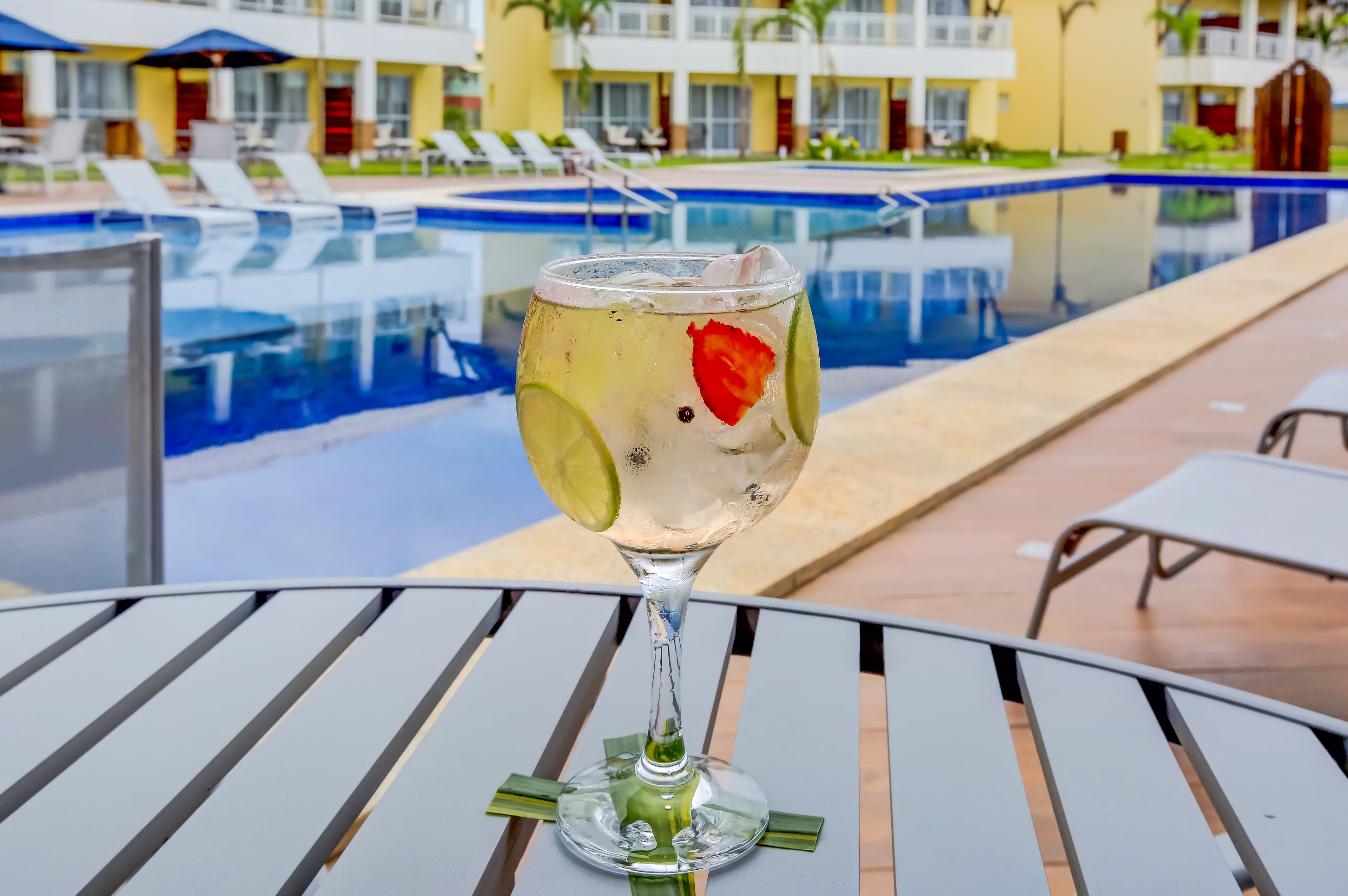 Un vaso de cóctel con lima y una fresa sobre una mesa, cerca de una piscina con sillas de descanso y un edificio de hotel al fondo.