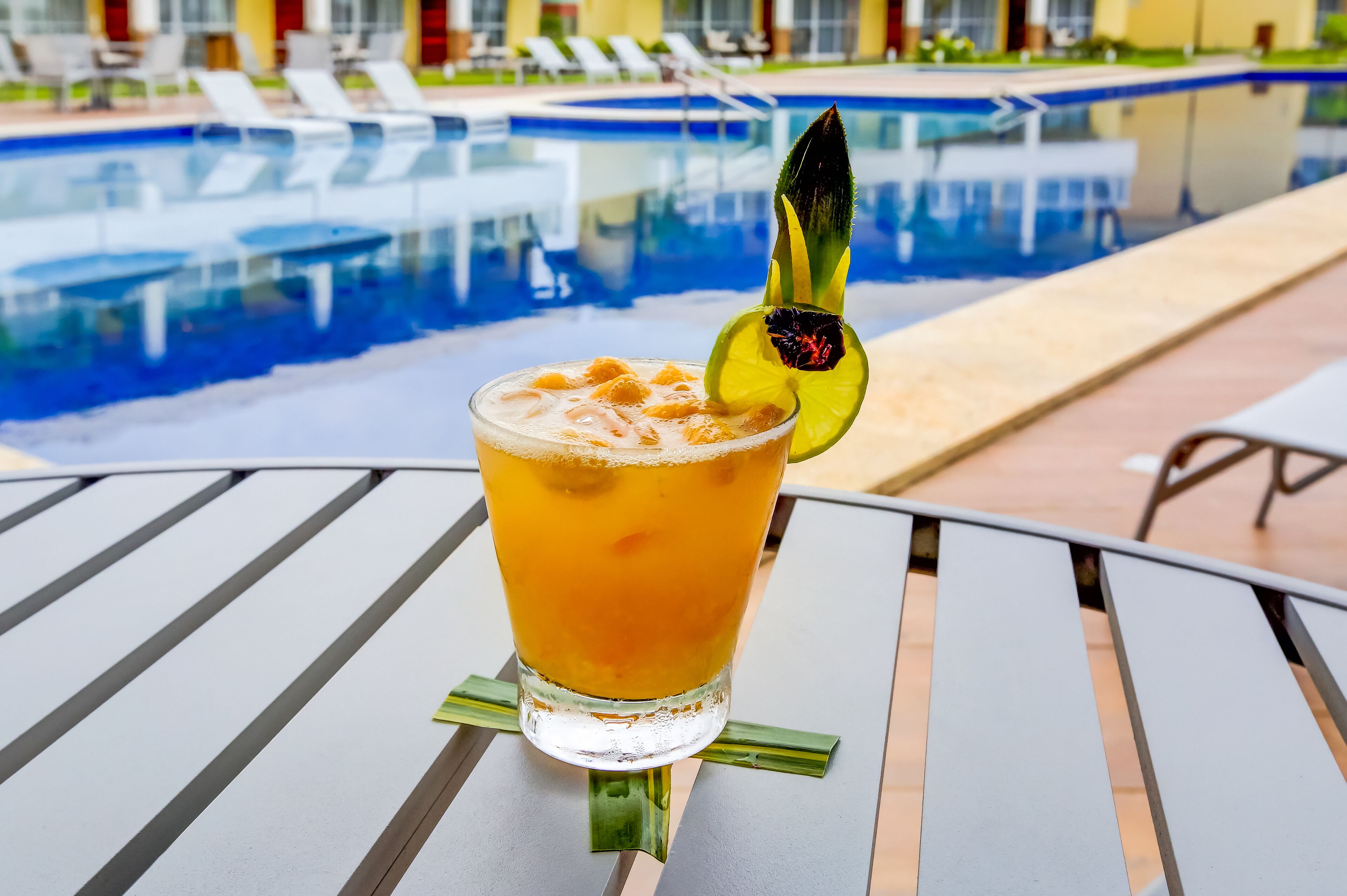 A glass of iced beverage with a lime garnish on a metal table by a poolside.