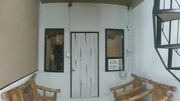 Ein Zimmer mit drei Holzstühlen, einem kleinen Tisch, einer weißen Tür und einem Fenster mit Metallrahmen.