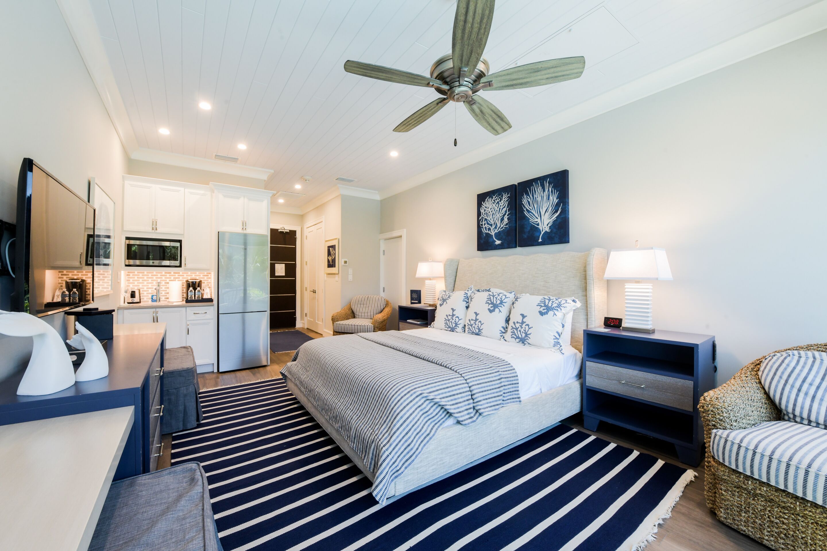 A modern bedroom with a bed, bedside tables, a nightstand, a chair, a ceiling fan, and a kitchen area in the background.