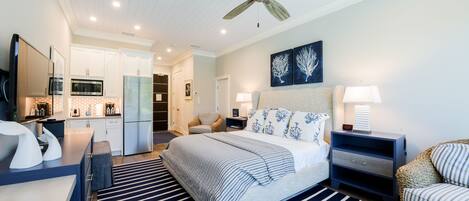 A modern bedroom with a bed, bedside tables, a nightstand, a chair, a ceiling fan, and a kitchen area in the background.