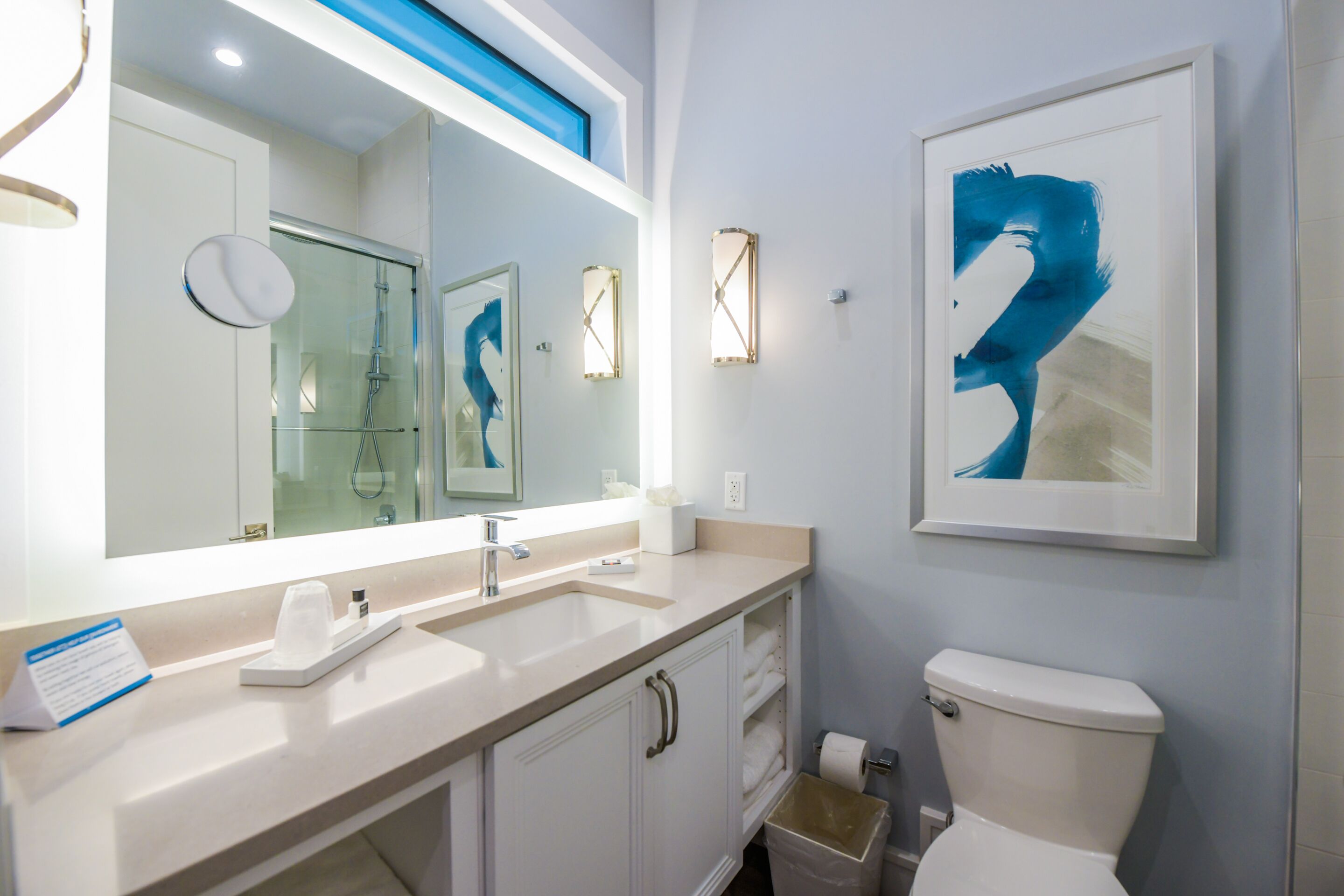 Un baño moderno con un espejo grande, un lavamanos, un inodoro y una obra de arte enmarcada en la pared.