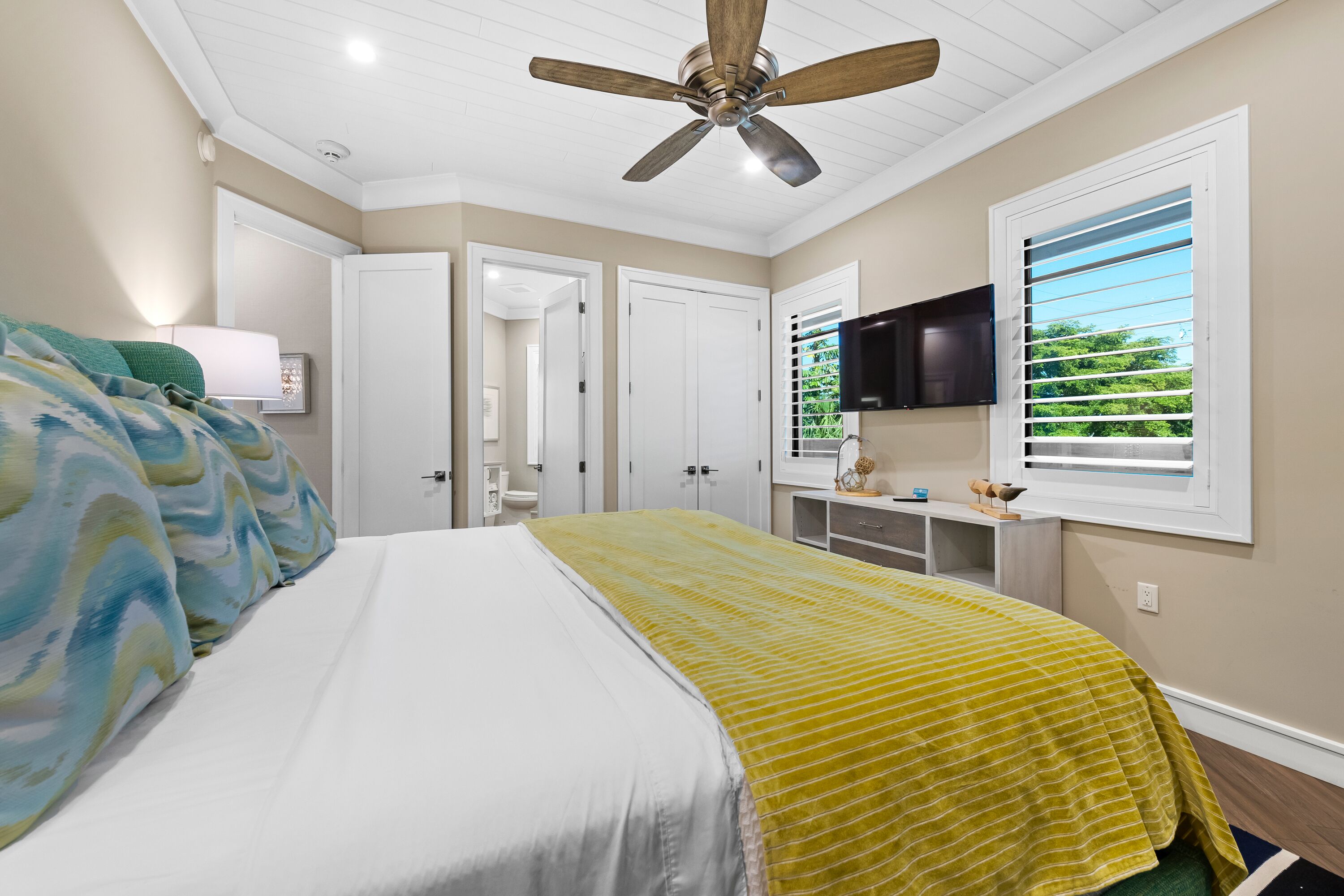 A bedroom with a bed, a ceiling fan, a TV, and a window with blinds.