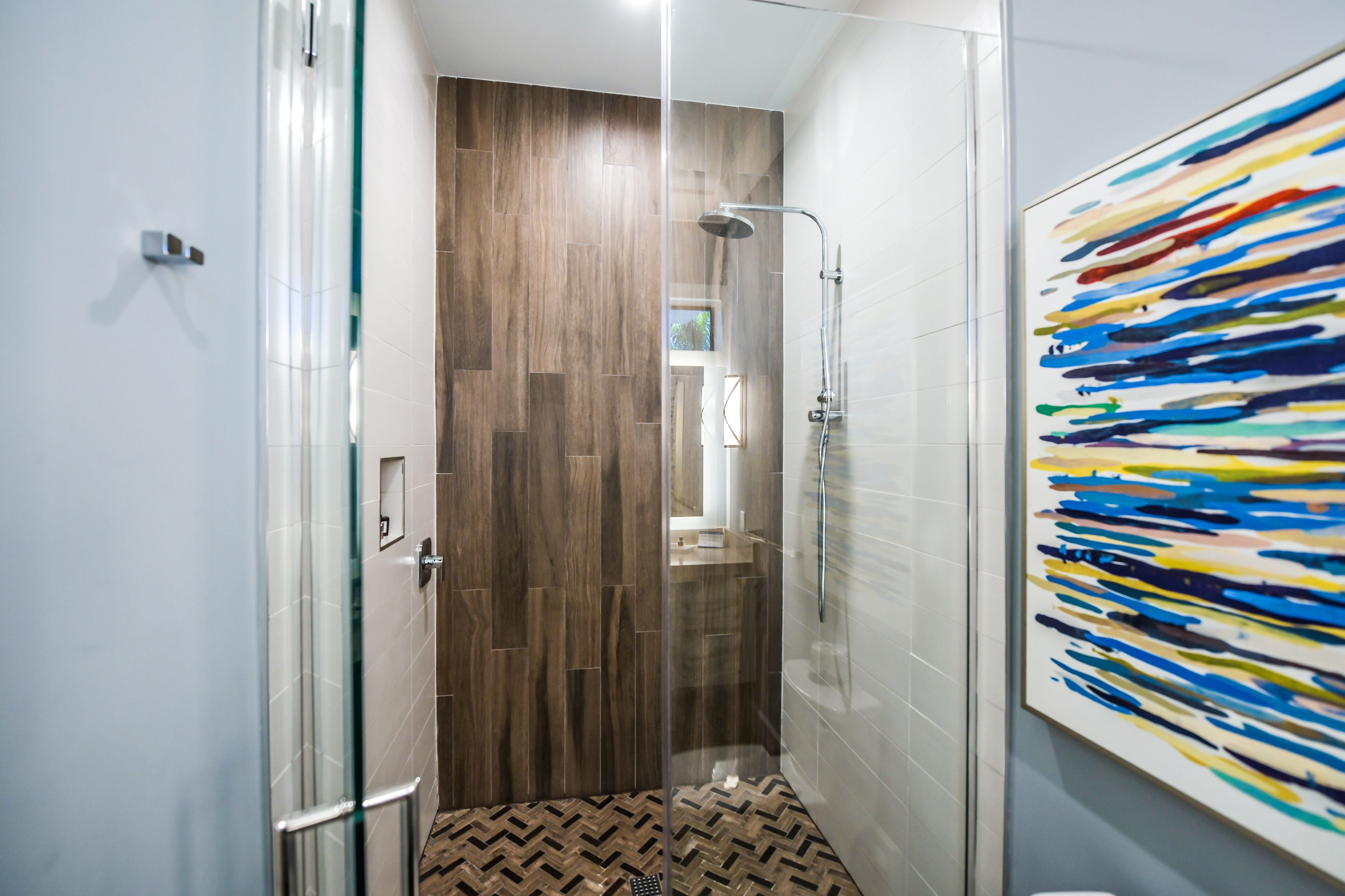 A modern bathroom with a glass shower enclosure, wooden flooring, and a large abstract painting on the wall.