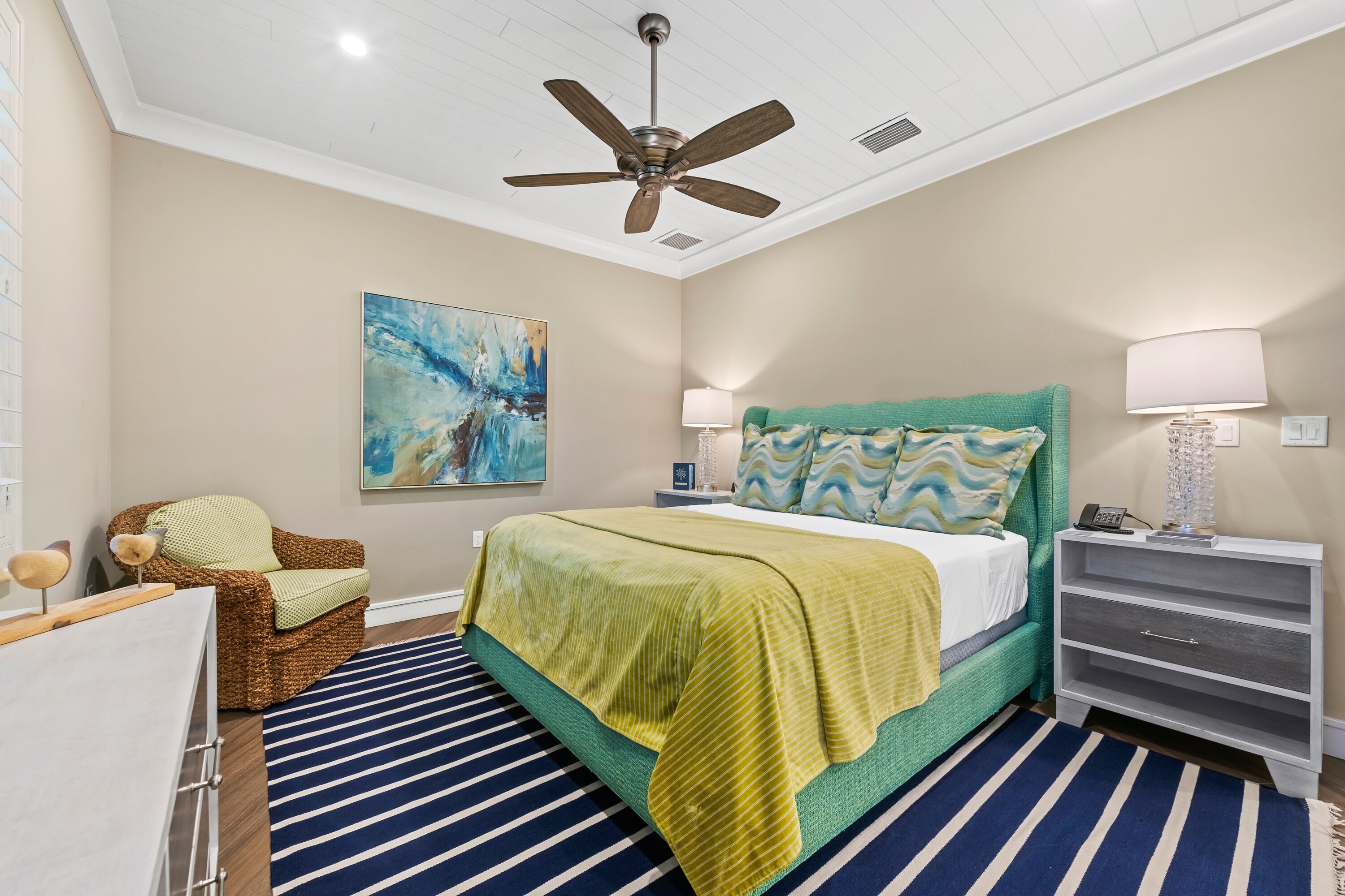 A bedroom with a bed, a ceiling fan, a nightstand, a lamp, a chair, and a painting on the wall.