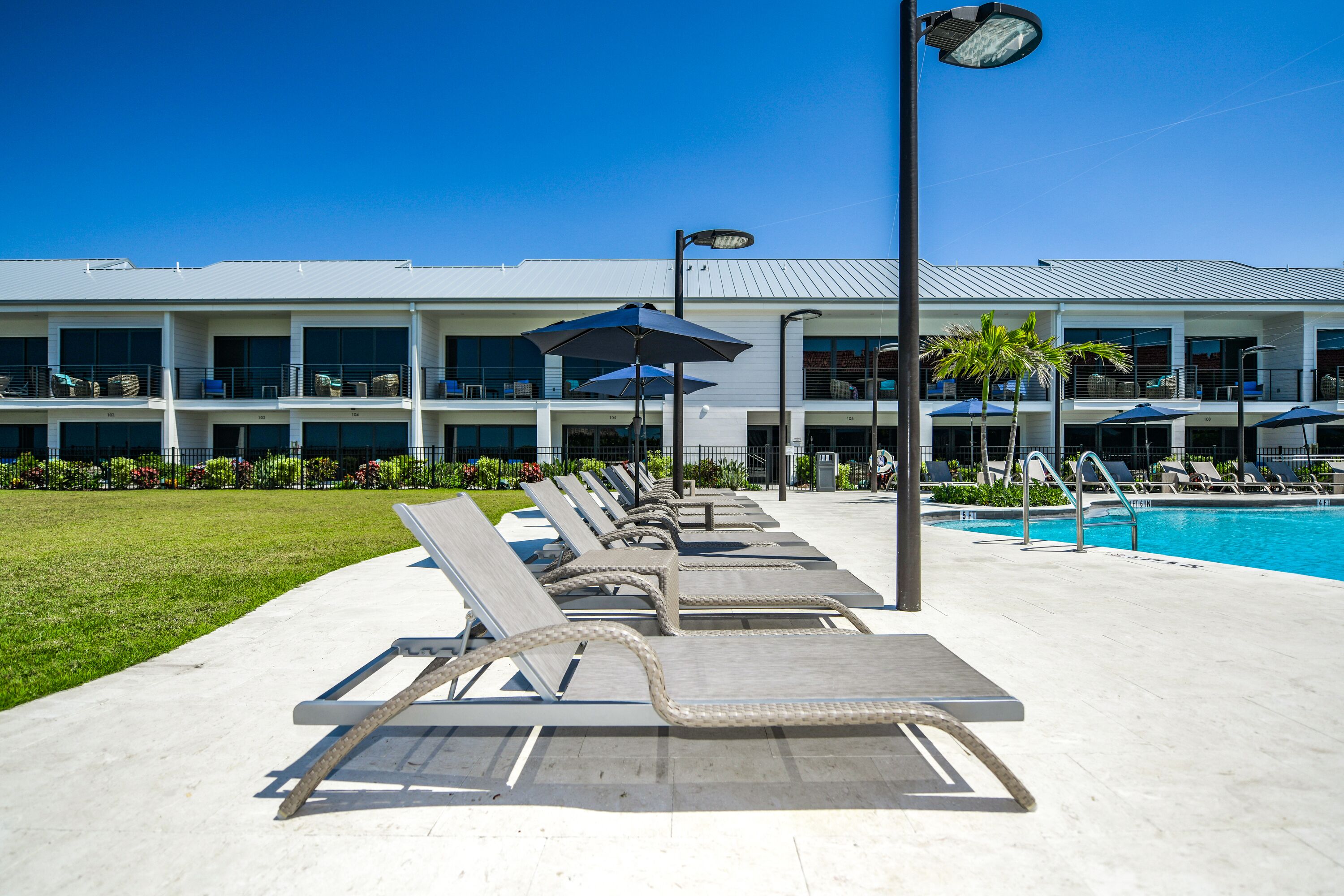 A poolside area with lounge chairs and umbrellas, a clear blue sky, and a building with multiple balconies in the background.