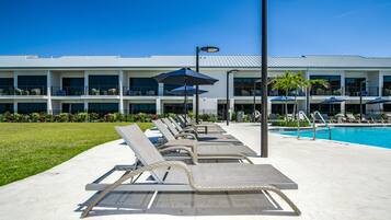 A poolside area with lounge chairs and umbrellas, a clear blue sky, and a building with multiple balconies in the background.