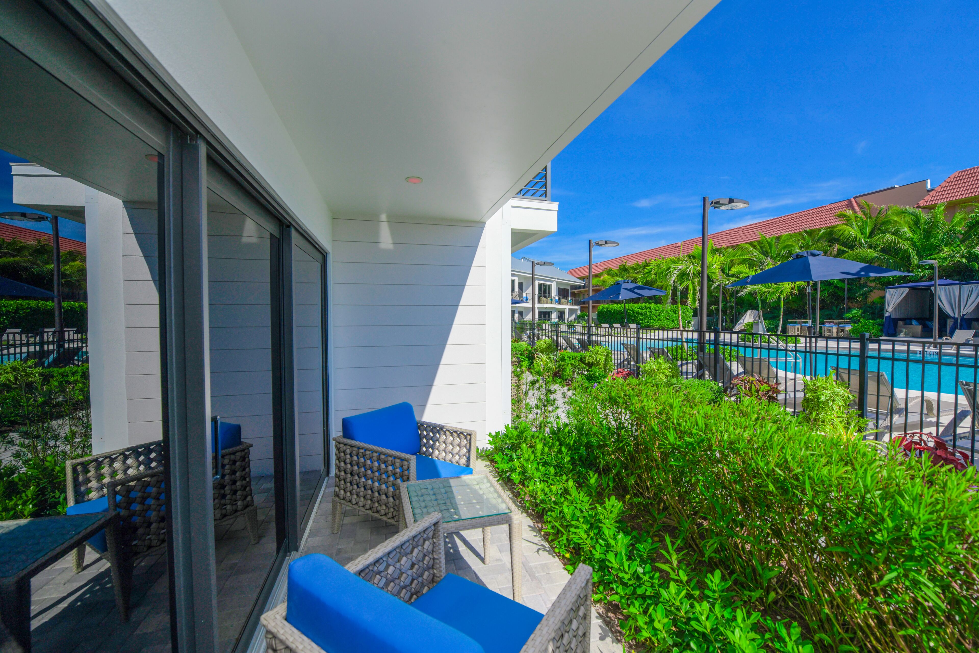 A balcony with wicker furniture and a glass table, overlooking a pool area with umbrellas and lounge chairs.