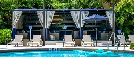 A pool area with lounge chairs, umbrellas, and a covered seating area with curtains.