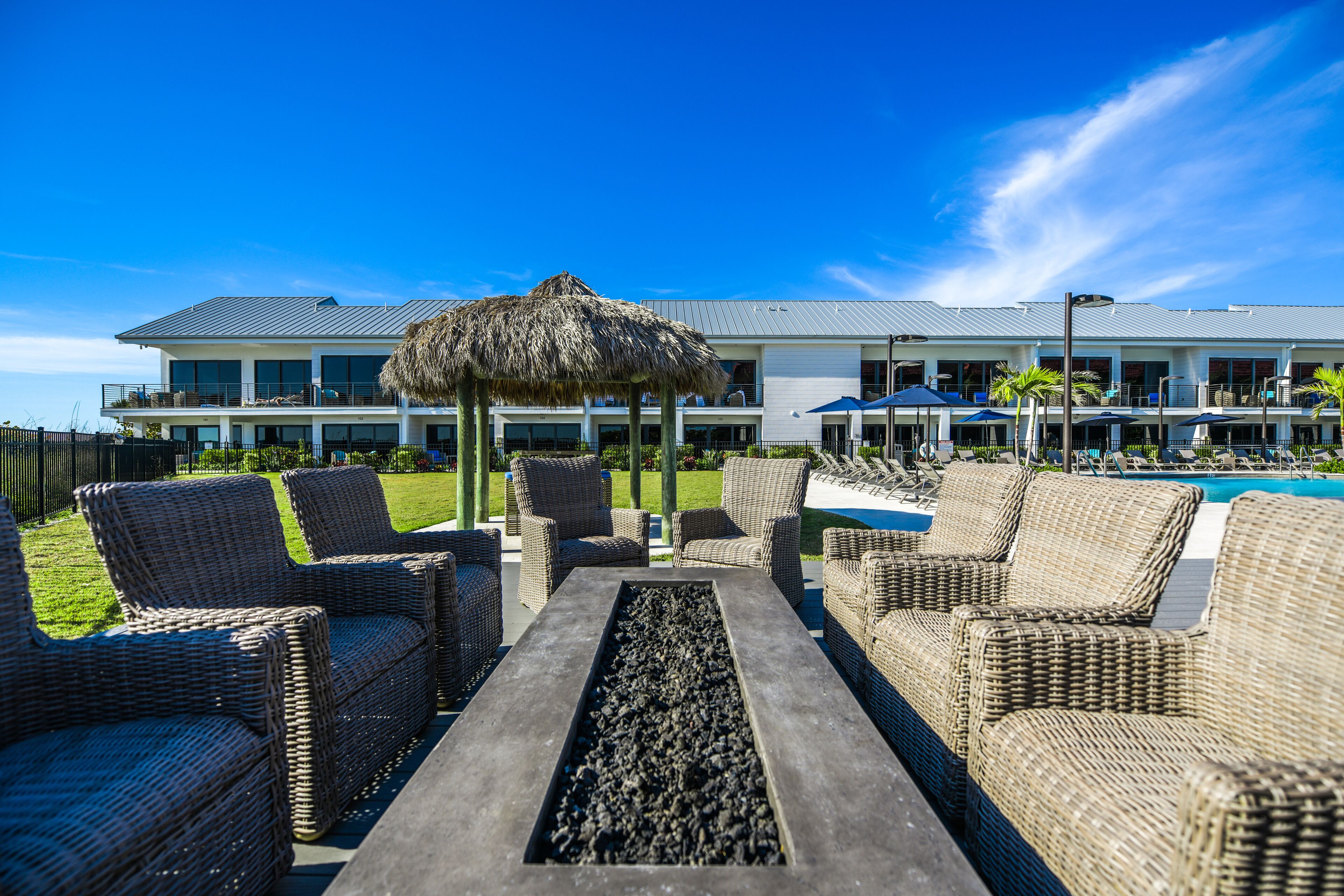 A poolside area with wicker furniture and a thatched umbrella, a pool, and a building with a modern design in the background.