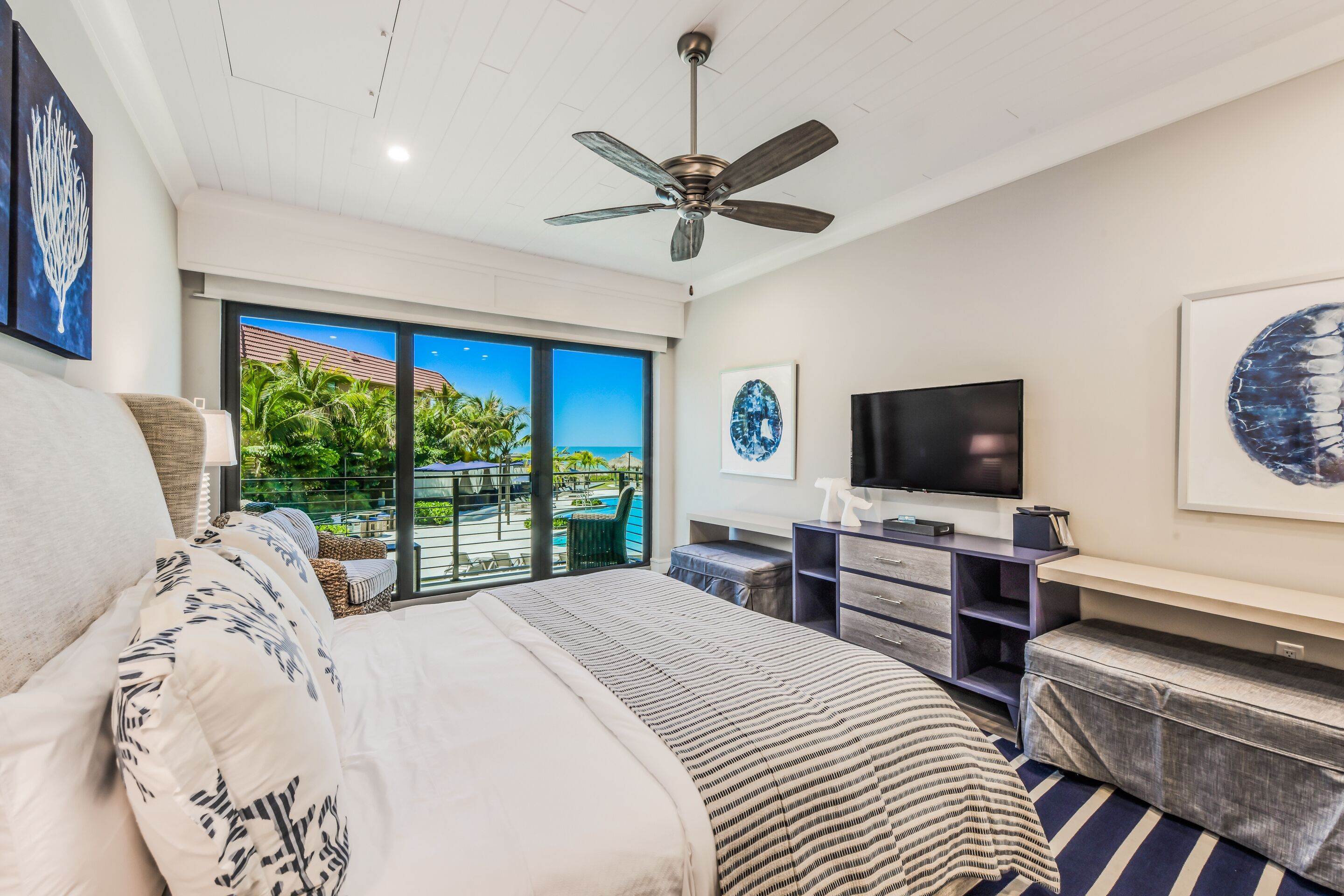 A modern bedroom with a large bed, a ceiling fan, a TV, and a balcony with a view of the ocean.