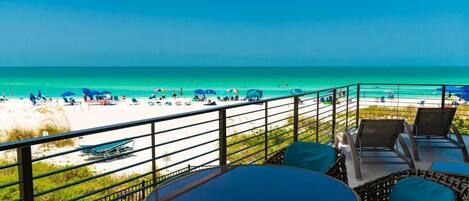 A balcony with wicker furniture and a table, overlooking a beach with umbrellas and people.
