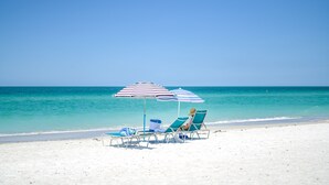 A beach with white sand, clear turquoise water, and a woman sitting under a striped beach umbrella. - Anna Maria Beach Resort (Holmes Beach)