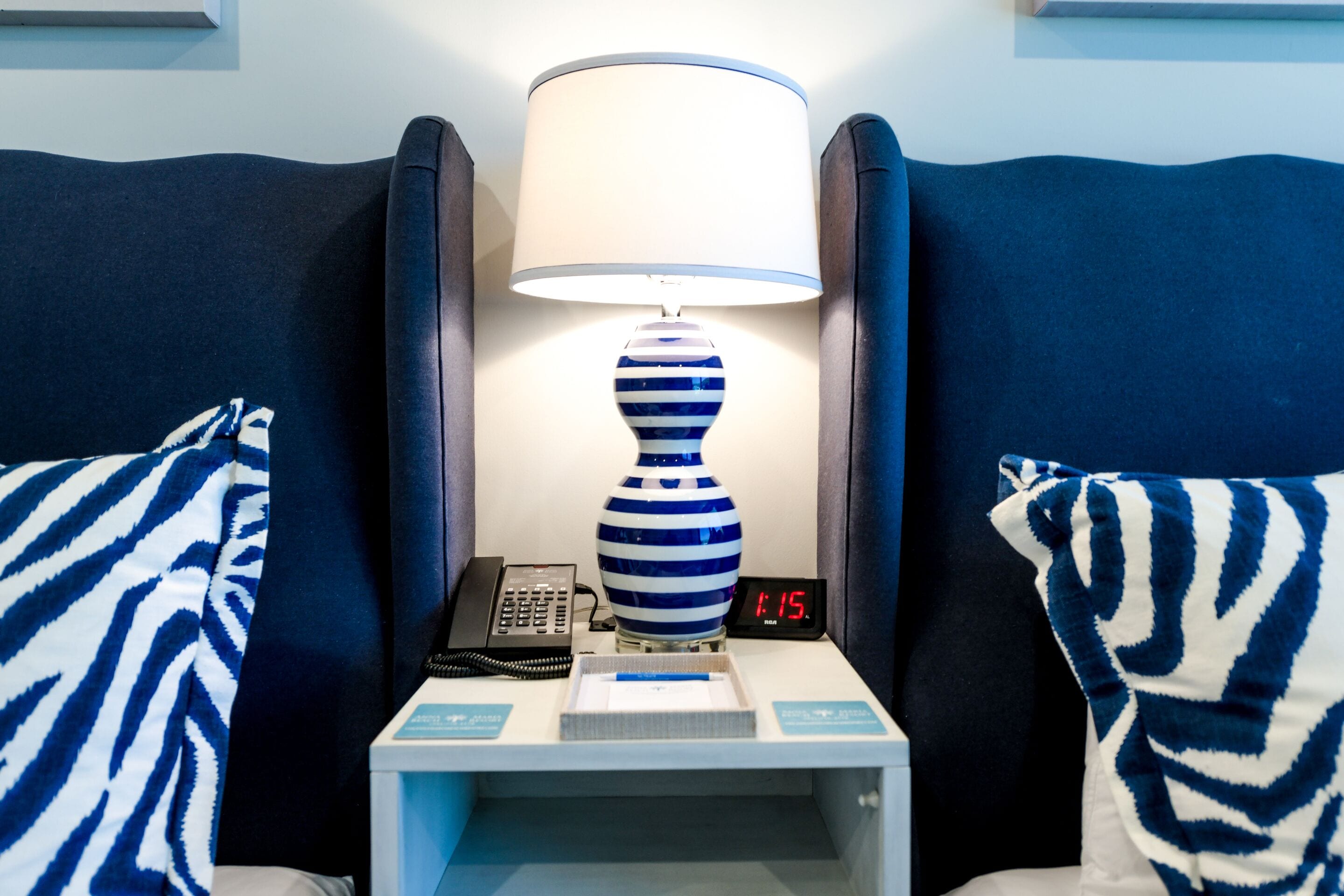 Une chambre d’hôtel avec une table de chevet sur laquelle se trouvent une lampe à rayures, un téléphone et un réveil numérique affichant 11h15.