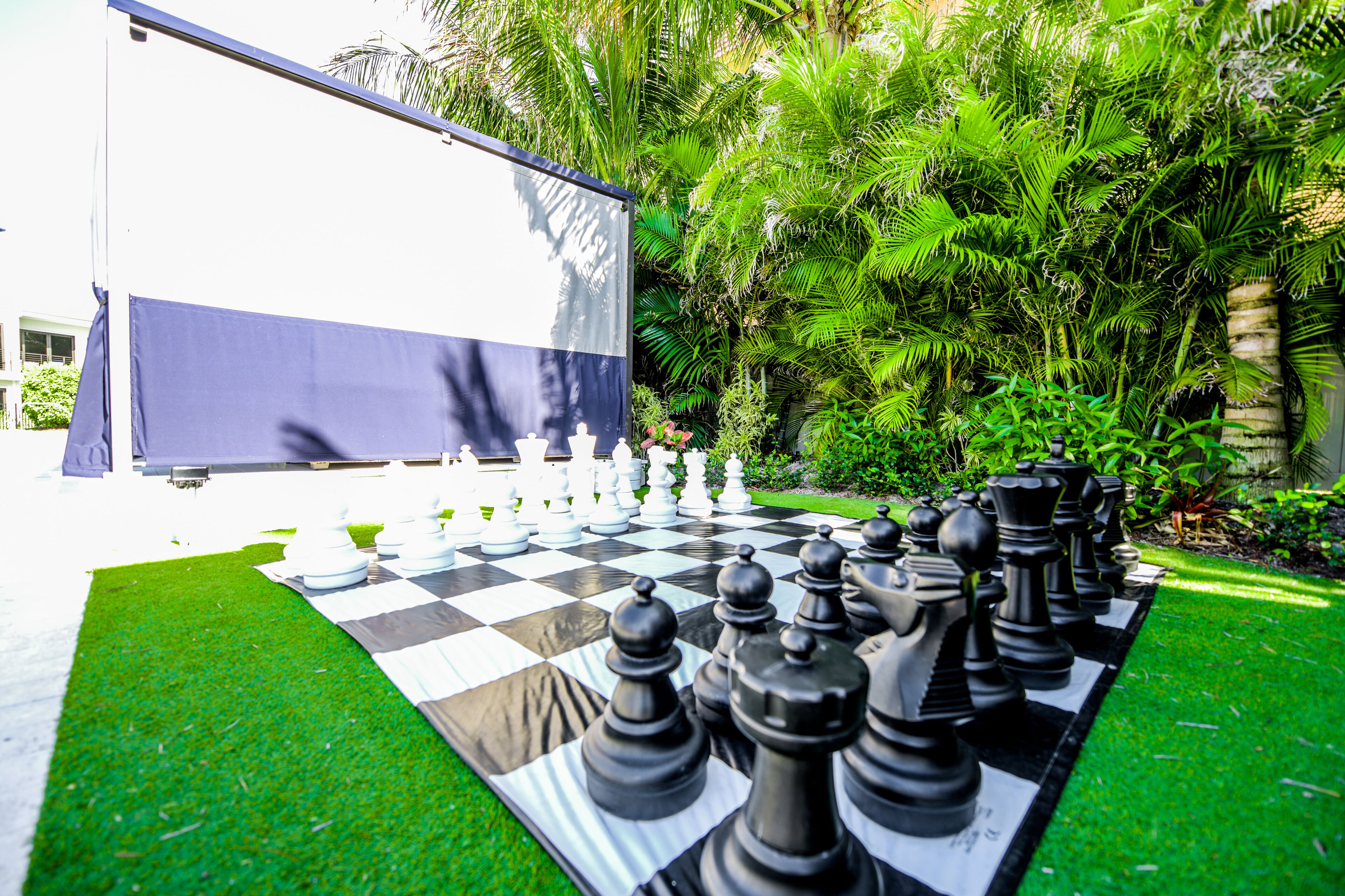 A large outdoor chessboard set up on artificial grass with a backdrop of dense tropical foliage.