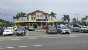 A building with a yellow facade and a red roof, surrounded by parked cars and palm trees. - Hotel O Z Dirgahayu 313 (Kota Bharu)