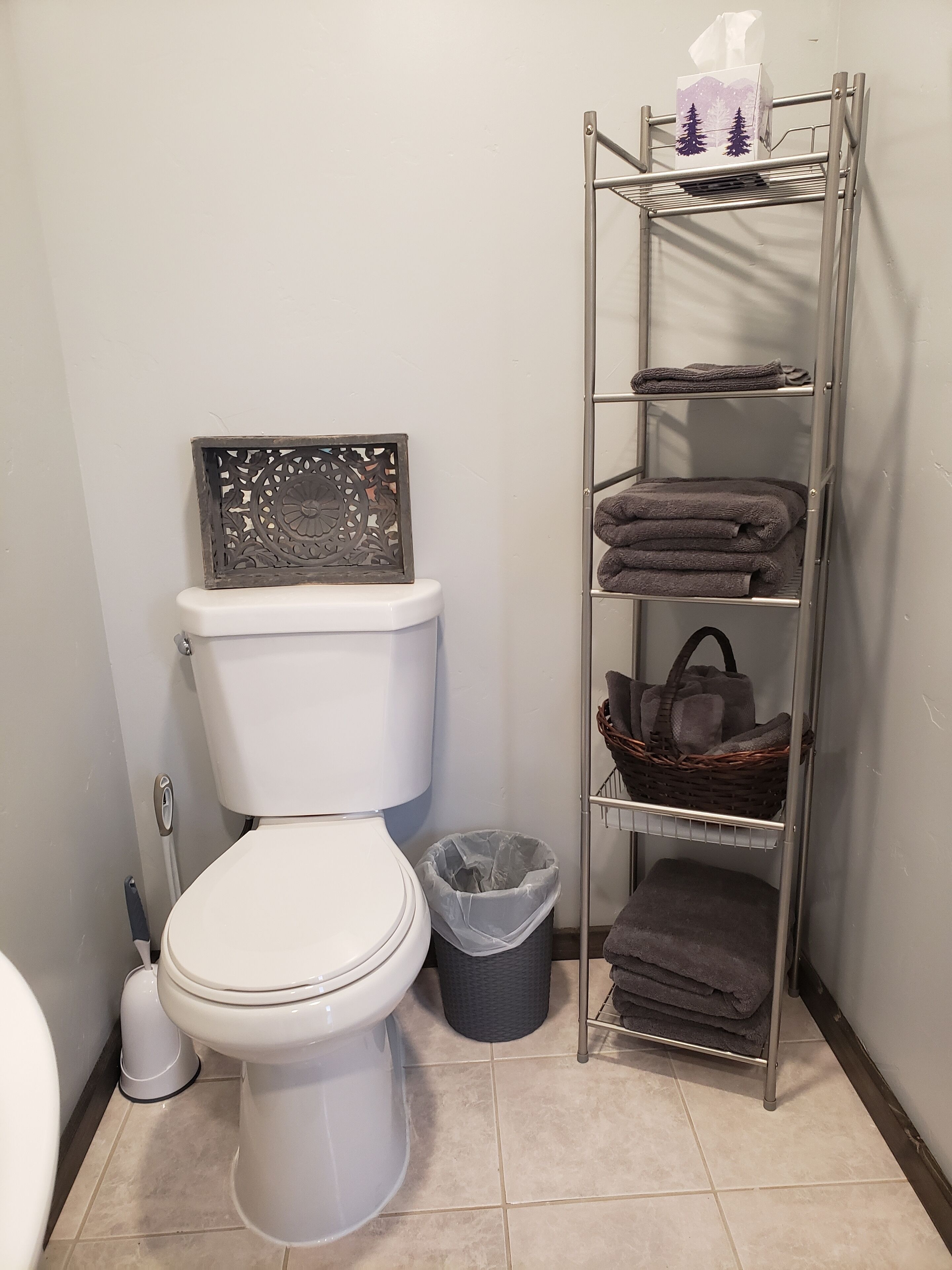 A bathroom with a toilet, a decorative wall piece, a shelf with towels and a basket, and a trash bin.