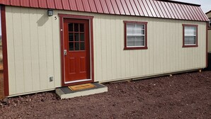 A single-story building with a red door and windows, a metal roof, and a number '5' on the wall. - LVBar Ranch - Cabin #2 (Cane Beds)