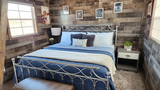 A bedroom with a metal bed frame, a wooden headboard, a bedside table with a lamp, and a small plant. - LVBar Ranch - Cabin #2 (Cane Beds)