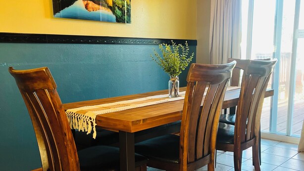 Un coin repas avec une table en bois, quatre chaises, un vase avec des plantes vertes et un tableau représentant un paysage accroché au mur.