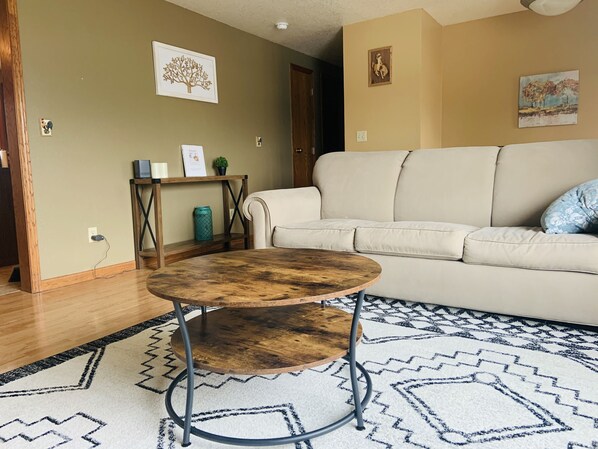 En stue med et rundt sofabord, en beige sofa, et mønstret tæppe og billeder på væggen.